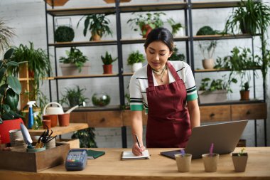 A beautiful Asian woman in an apron works in her plant shop, taking notes while managing her small business. clipart