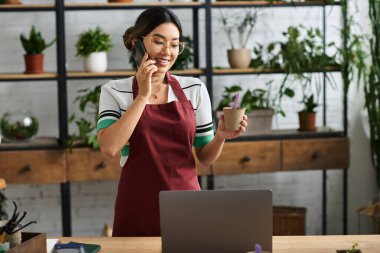 Önlüklü gülümseyen Asyalı bir kadın, küçük bir fabrika sahibi, elinde saksı bitkisi tutarken telefonla konuşuyor..
