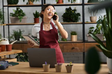 Önlüklü mutlu bir Asyalı kadın çiçekçi dükkanında duruyor ve telefonuna telefon açıyor..