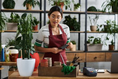 An Asian woman in an apron stands behind the counter of her plant shop clipart