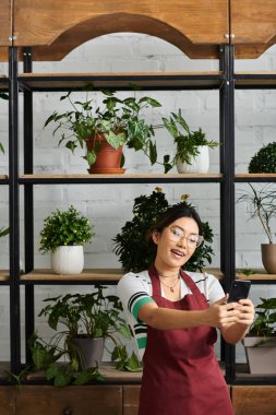 Önlüklü mutlu bir Asyalı kadın fabrikada çalışıyor. Telefonunu kullanırken kameraya gülümsüyor..