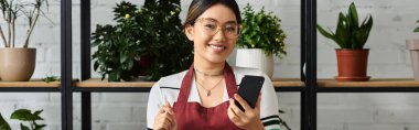 Gülümseyen Asyalı bir kadın, çiçeklerle ve yeşilliklerle çevriliyken telefonunu kullanıyor..