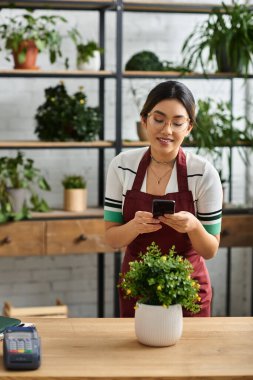 A smiling Asian woman, wearing an apron, works at the counter of her plant store, checking her phone. clipart