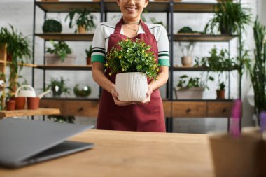 A smiling Asian woman wearing an apron holds a potted plant in her plant shop. clipart