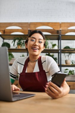 An Asian woman in an apron smiles while working on a laptop in her plant shop. clipart