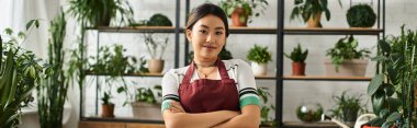 A smiling Asian woman in an apron stands confidently in her plant shop, surrounded by lush greenery. clipart