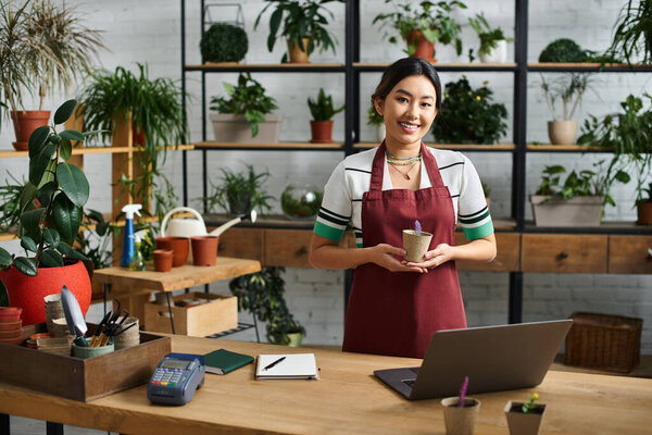 An Asian woman, the owner of a plant store, smiles as she holds a small potted plant.