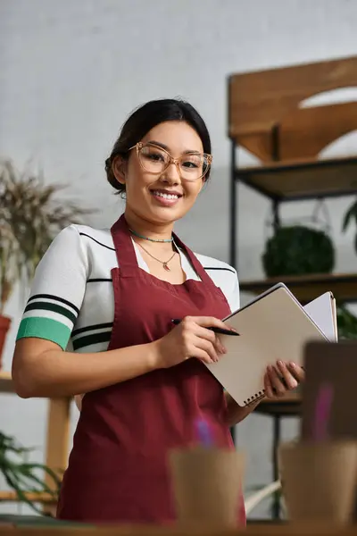 stock image A young Asian woman in an apron stands in her plant shop, smiling and holding a notepad.