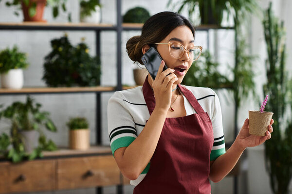 A young woman in a red apron talks on her phone while holding a potted plant in her other hand.