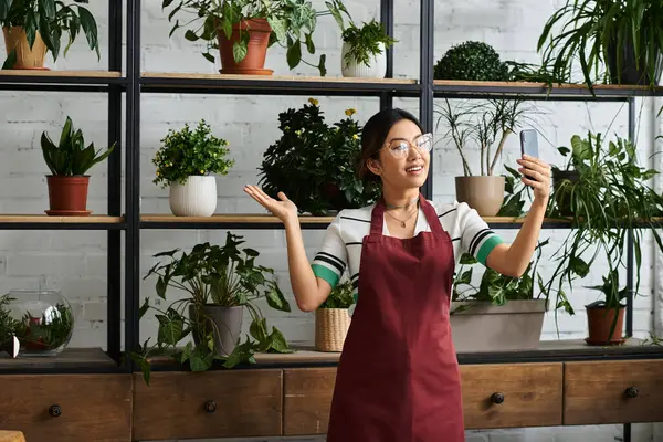 Kırmızı önlüklü gülümseyen Asyalı bir kadın yemyeşil çiçeklerle çevrili bitki atölyesinde selfie çekiyor..
