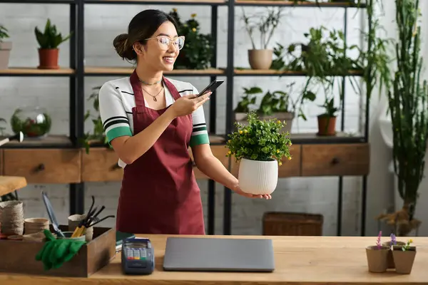 Bir kadın çiçekçisinde saksı bitkisini tutarken gülümsüyor. Telefonda konuşuyor gibi görünüyor, muhtemelen sipariş alıyordur..