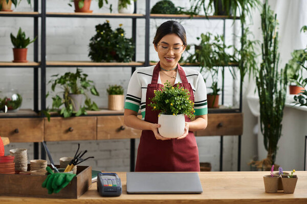 A smiling Asian woman in an apron stands in her plant shop, holding a potted plant.