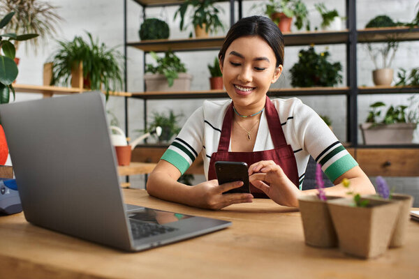 A smiling Asian woman, wearing an apron, checks her phone while working in her plant store.