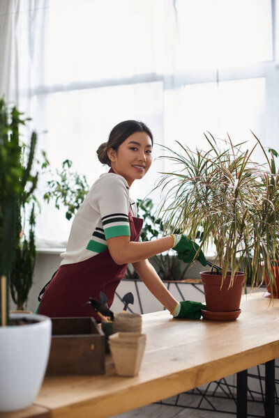 A young Asian woman, wearing an apron, smiles while tending to plants in her flower shop.
