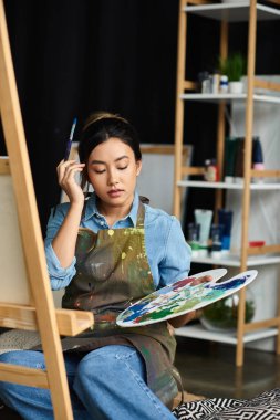 A young Asian woman sits in her art studio, holding a paintbrush and palette, focused on her artwork. clipart