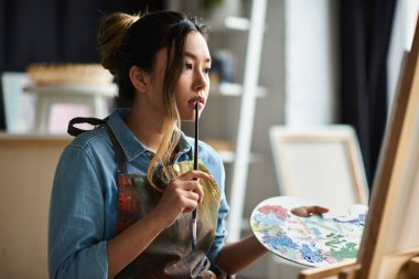 A young Asian woman, wearing an apron, holds a paint palette and contemplates her next artistic move in her workshop. clipart