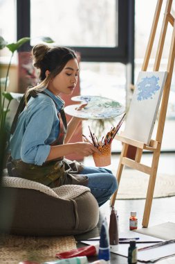 A young Asian woman in an apron sits on a cushion in her workshop, holding a paintbrush and concentrating on her painting. clipart