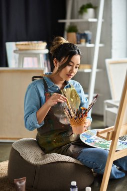 An Asian artist, wearing an apron, sits in her workshop and focuses on her painting. clipart