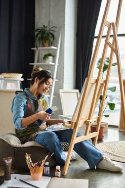Genç bir Asyalı sanatçı atölyesinde oturuyor, resim yapmaya odaklanmış, etrafı sanat malzemeleri ve doğal ışıkla çevrili..