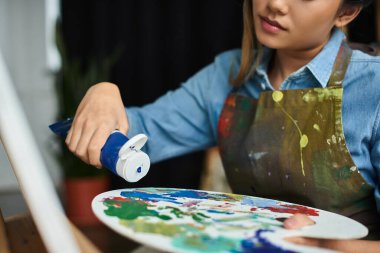 A young Asian woman in an apron squeezes paint from a tube into a palette in her workshop. clipart