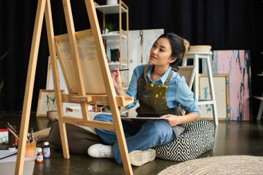 A young Asian woman in an apron paints on a canvas in her workshop. clipart