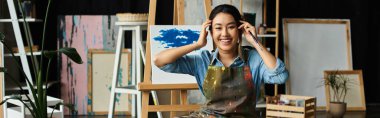 A young Asian artist, wearing an apron, smiles brightly as she takes a break from painting in her workshop. clipart