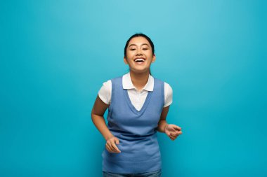 A young Asian woman wearing a white shirt and blue vest smiles brightly against a blue backdrop. clipart