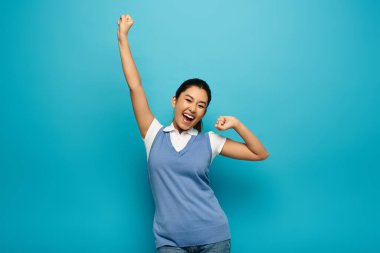 Asian woman in smart casual outfit, smiling and celebrating with raised arms against blue background. clipart