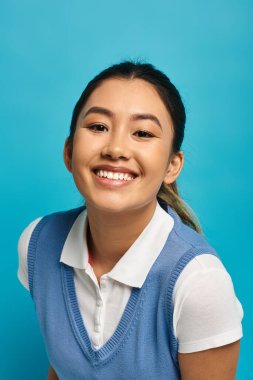 Beyaz gömlek ve mavi yelek giyen genç bir Asyalı kadın mavi arka plana doğru gülümsüyor..