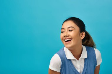 Zeki, sıradan giyinmiş genç bir Asyalı kadın mavi bir arka plana parlak bir şekilde gülümsüyor. İfadesi gerçek neşe ve mutluluk veriyor..