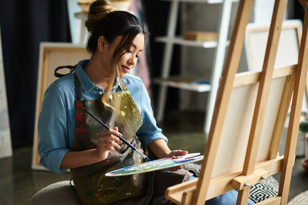 Молодая азиатка в фартуке сидит в своей мастерской, сосредоточенная на смешивании краски на своей палитре.