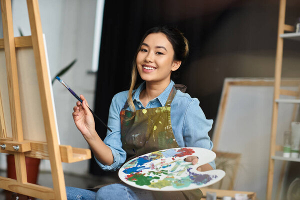 Счастливая азиатская художница рисует в своей мастерской, держа кисть и палитру.