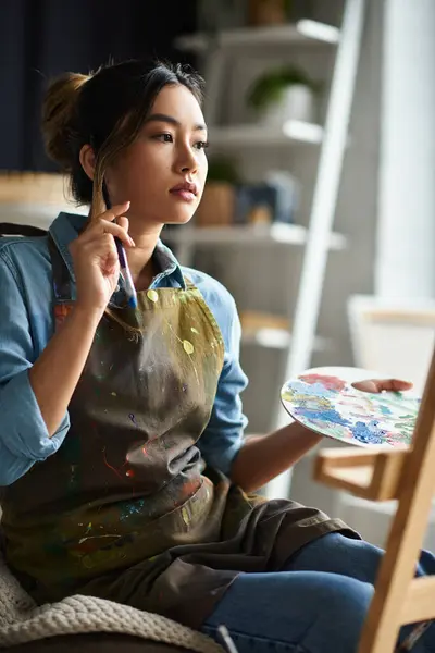 stock image Asian female artist in paint-splattered apron in workshop, holding a brush, contemplating next stroke.