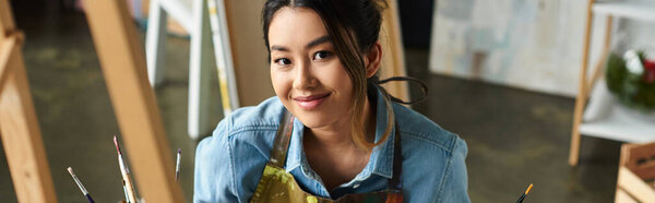 Молодая азиатка, одетая в фартук, улыбается, когда работает над картиной в своей художественной студии.