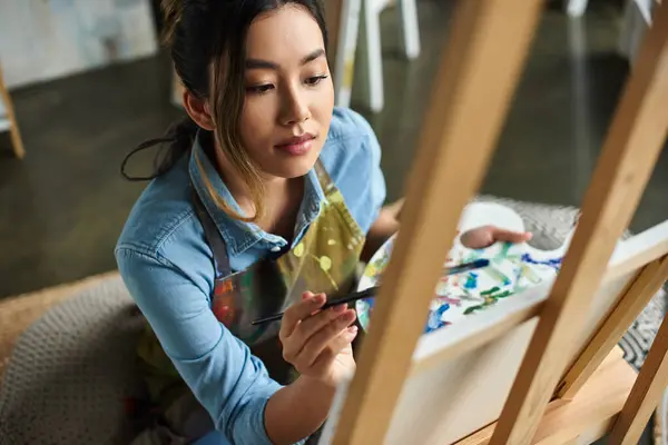 Stock image A young Asian artist, wearing an apron, paints on a canvas in her workshop.