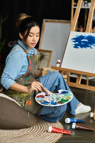 A young Asian woman wearing an apron sits in her workshop and mixes paints on a palette.