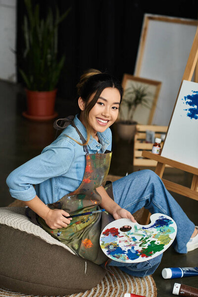A young Asian artist in a workshop, wearing an apron, smiles while holding a paintbrush and palette.