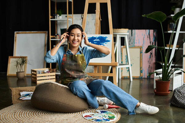 Молодая азиатка в фартуке сидит на кресле-мешке в своей художественной студии, улыбаясь и держа кисть.