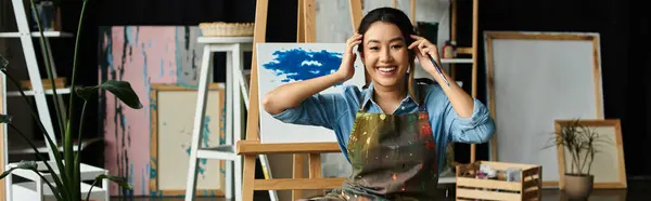 stock image A young Asian artist, wearing an apron, smiles brightly as she takes a break from painting in her workshop.