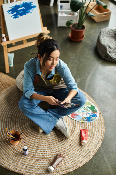 A young Asian woman sits on a round rug in her workshop, carefully choosing paint colors from her palette.
