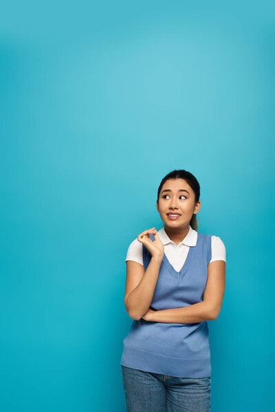 Молодая азиатка в умной повседневной одежде улыбается и смотрит в сторону, стоя на синем фоне.