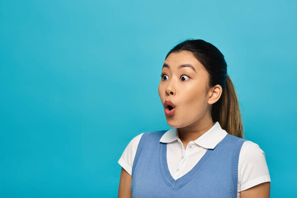 A young Asian woman in smart casual attire stands against a blue background, looking surprised and with her mouth open.