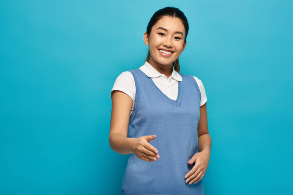 A young Asian woman in a smart casual outfit smiles and offers her hand for a handshake against a bright blue background.