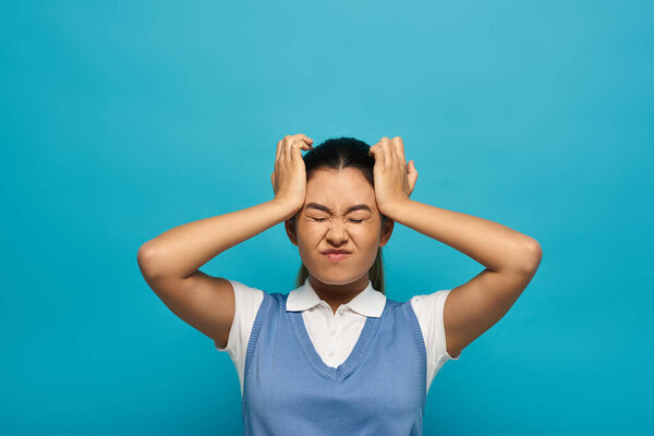Разочарованная молодая женщина в умной повседневной одежде с темными волосами и голубым топом держит голову на синем фоне.