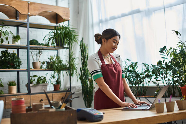 A smiling Asian woman wearing an apron works on laptop in her plant store, surrounded by greenery.