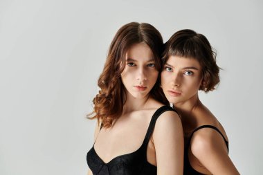 Siyah elbiseli iki genç kadın gri bir arka plana karşı birlikte poz veriyorlar..