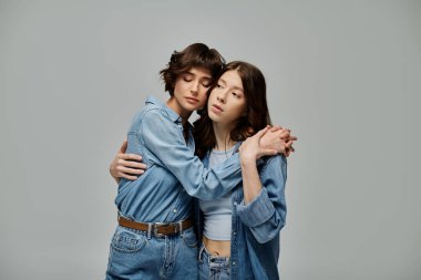 Kot giymiş iki genç kadın, gri bir arka planda birbirlerine sarılıyorlar..