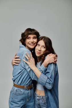 Kot pantolon giymiş iki şık genç kadın gri bir arka plana karşı poz veriyor, birbirlerini sevinçle kucaklıyorlar. Lgbt çifti