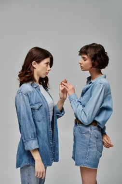 Kot gömlekli ve kot pantolonlu iki genç kadın gri bir arkaplanda poz veriyor..