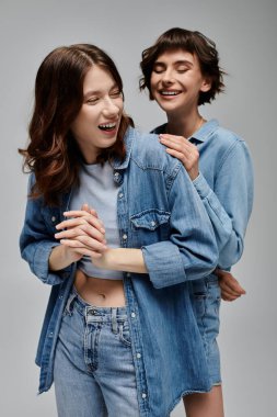 Kot pantolon giymiş iki genç kadın gri bir arkaplan önünde birlikte gülüyorlar. Lezbiyen çift